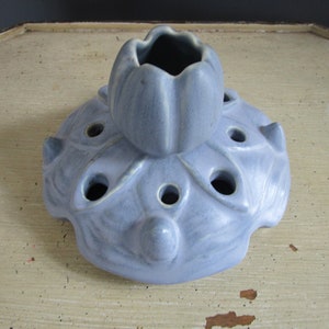May include: A light blue ceramic flower frog with a tulip-shaped top and multiple holes for arranging flowers.
