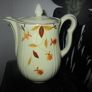 May include: A white ceramic coffee pot with a brown and yellow floral pattern. The pot has a lid with a knob on top and a handle on the side.