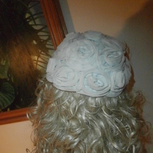 May include: A light blue hat with a design of multiple fabric rosettes. The hat is on a mannequin with blonde curly hair.