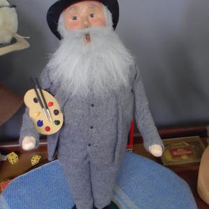 May include: A figurine of an elderly person with a long white beard wearing a black hat and a grey suit. The figure holds a wooden artist palette with colorful paint dabs and two thin brushes in one hand, standing on a circular green base.