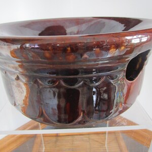 May include: A brown glazed ceramic planter with a scalloped rim and a drainage hole. The planter has a glossy finish and is decorated with a raised design.
