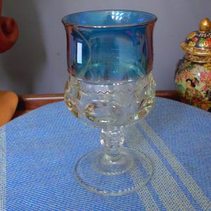 May include: A clear glass goblet featuring a wide, iridescent blue upper bowl and a textured lower section with a repeating circular pattern. The stem is short and clear, leading to a circular base, creating a distinct two-tone appearance.