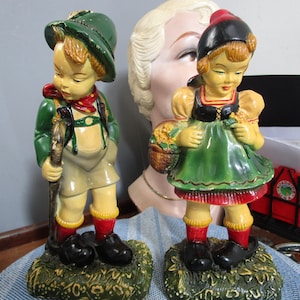 May include: Two vintage ceramic figurines of children in traditional German attire. The boy wears a green hat and jacket, holding a walking stick. The girl has a black hat and green dress, holding a basket. Both stand on a green base.