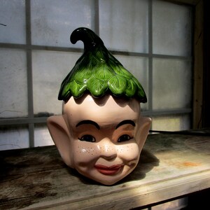 May include: A ceramic figurine of a gnome with a green leaf hat. The gnome has a white face with brown eyes and a red mouth. The gnome is wearing a white beard.