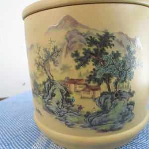 May include: A cylindrical ceramic container with a beige background and a painted scene of a mountain landscape with trees, houses, and a river. The scene is painted in shades of green, blue, and brown.