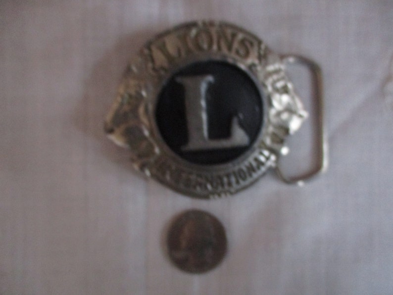 Puede incluir: Hebilla de cintur&oacute;n de plata con un dise&ntilde;o en blanco y negro. La hebilla tiene el texto "LIONS INTERNATIONAL" y una gran letra "L" en el centro.