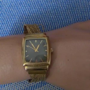 May include: A gold-toned rectangular wristwatch with a dark face and gold hands. The watch has a gold mesh band. The watch is on a wrist, against a blue and white striped background.