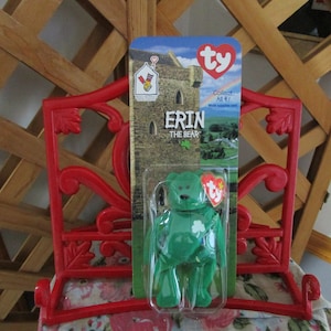 May include: A green plush bear with a shamrock on its chest, labeled "Erin the Bear" on a Ty Beanie Baby packaging card. The card features a castle and a rainbow.