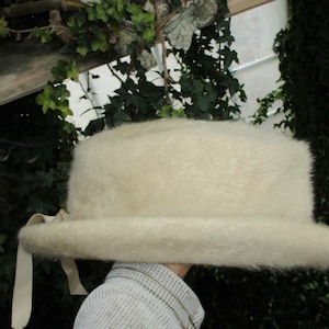 May include: A cream-colored, fuzzy hat with a wide brim and a ribbon detail. The hat has a cylindrical crown and a flat, rounded brim. The hat is made of a soft, textured material.