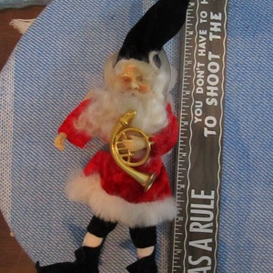 May include: A red and white elf figurine with a black hat and a white beard, holding a gold trumpet. The elf is standing on a blue and white patterned surface. A wooden ruler with the text "As a rule you don't have to hunt to shoot the bull" is visible on the right side of the image.