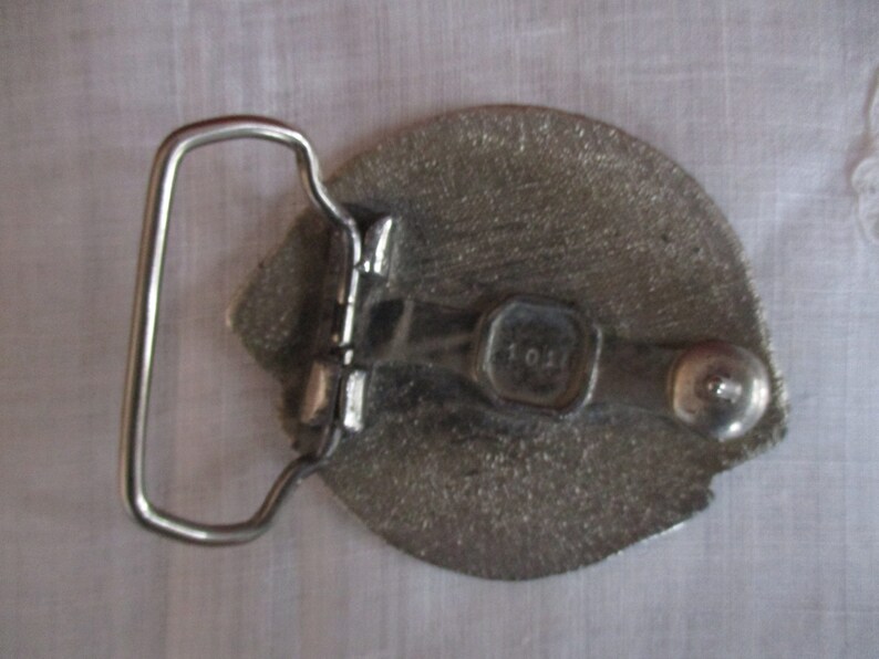 Puede incluir: Hebilla de cintur&oacute;n de plata con un dise&ntilde;o detallado y un lazo de plata. La hebilla tiene una peque&ntilde;a etiqueta de metal con el n&uacute;mero "101" en ella.