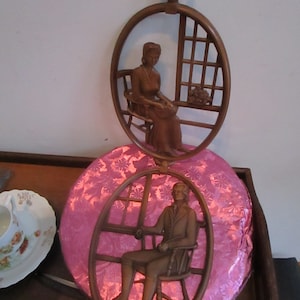 May include: A pair of wooden wall hangings depicting a man and woman sitting in chairs. The hangings are circular and have a pink background.