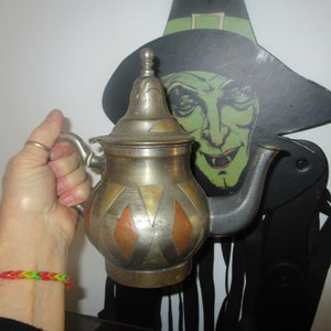 May include: A vintage metal teapot with a lid, handle, and spout. The teapot has a silver finish with gold and copper diamond-shaped accents. A Halloween witch decoration is in the background.