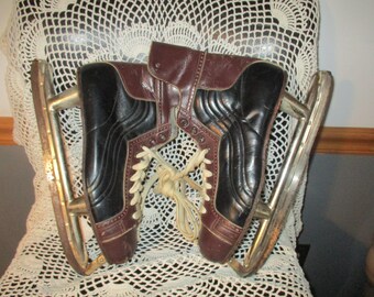 May include: A pair of vintage ice skates with black leather uppers and brown leather accents. The skates have white laces and silver metal blades.