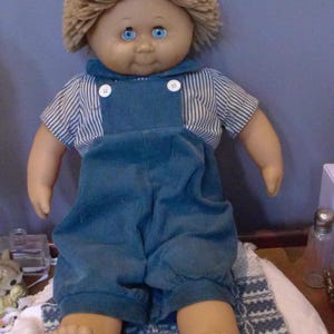 May include: A vintage doll with a tan face, blue eyes, and light brown yarn hair. The doll wears a blue corduroy overall and a striped shirt. The doll is sitting on a patterned blanket.