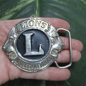 Puede incluir: Hebilla de cintur&oacute;n de plata con una cabeza de le&oacute;n negra y plateada a cada lado de un c&iacute;rculo con las palabras "Lions International" y una gran "L" plateada en el centro.