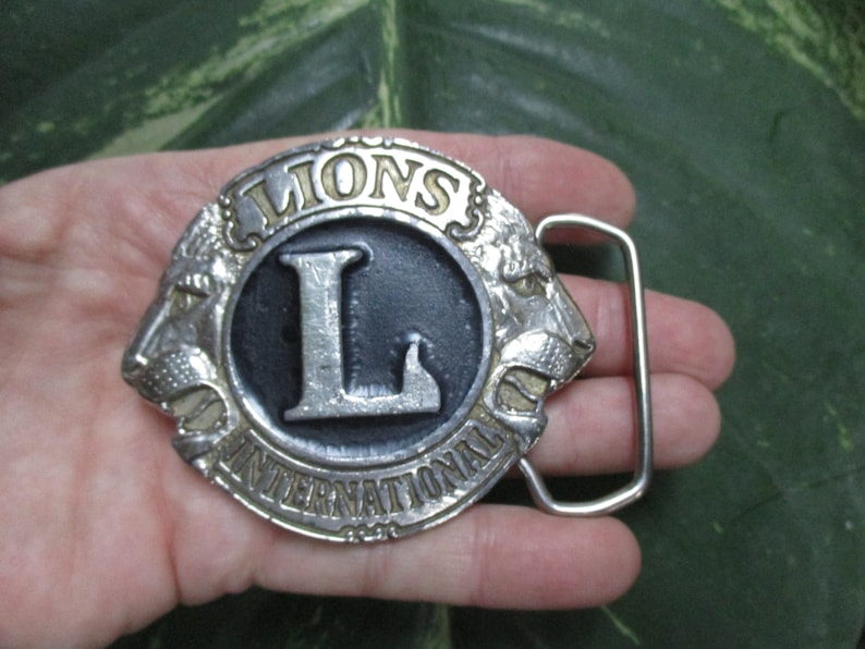 Puede incluir: Hebilla de cintur&oacute;n de plata con una cabeza de le&oacute;n negra y plateada a cada lado de un c&iacute;rculo con las palabras "Lions International" y una gran "L" plateada en el centro.