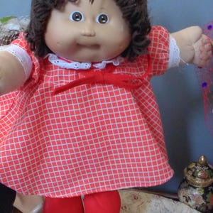 May include: A vintage Cabbage Patch Kid doll wearing a red and white checkered dress with a white lace collar and cuffs. The doll has brown yarn hair and red leggings. The doll's face is pale with large blue eyes.