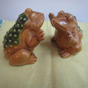 May include: Two ceramic frog figurines. One frog has a green and brown textured back, the other is smooth. Both are in a light brown color with glossy finish. The frogs are posed as if they are interacting with each other.