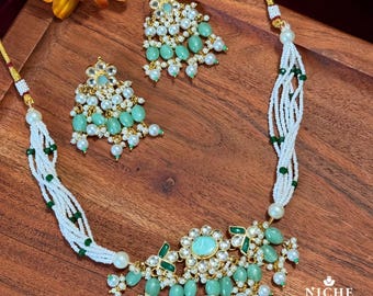 Op de afbeelding: Een witte en groene kralenketting met een grote groene en gouden hanger. De hanger is versierd met witte stenen en heeft een bloemmotief. De ketting wordt geleverd met bijpassende oorbellen.