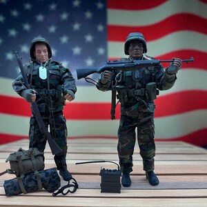 May include: Two action figures dressed in military camouflage uniforms, helmets, and boots, holding rifles. One figure has a backpack and a camouflage-patterned bag. A black radio and a pair of goggles are on the wooden surface. The American flag is in the background.