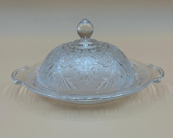 May include: A clear glass butter dish with a domed lid and a decorative floral pattern. The dish has a round base with handles and a knob on top of the lid. The glass is transparent, allowing the contents to be visible.