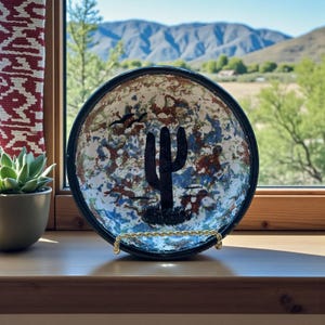 May include: A decorative ceramic bowl with a black rim and a painted desert scene. The bowl features a black cactus, birds, and a colorful splatter design in blue, red, and white. It sits on a gold-colored stand.