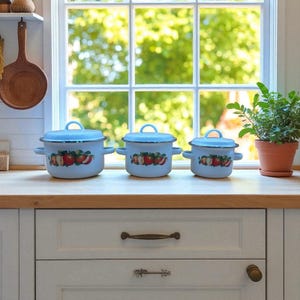 May include: Three light blue enamel pots with lids and handles, arranged on a wooden countertop. Each pot features a decorative band of red apples and green leaves. The pots are of varying sizes, suitable for cooking.