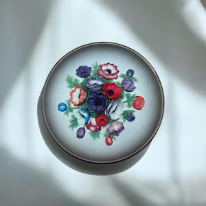 May include: A ceramic plate with a raised floral design. The plate features a bouquet of colorful anemones in shades of red, pink, purple, blue, and white. The flowers are surrounded by green leaves and are set against a light gray background.