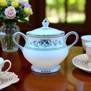 以下が含まれることがあります： 白磁の砂糖入れ、蓋付き、青、黄、ピンクの花柄。ボウルには2つのハンドルとシルバーのトリムが付いています。背景には花瓶に入った花があります。