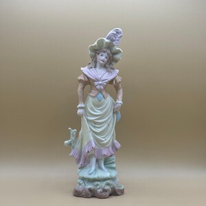 May include: A porcelain figurine of a woman in a yellow dress with pink accents, wearing a hat with a feather. The figurine is standing on a green and brown base.
