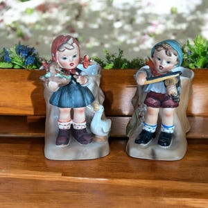 May include: Two vintage ceramic figurines. One is a girl in a blue dress holding a basket, with a white duck figurine. The other is a boy playing a violin. Both are in front of a white vase.