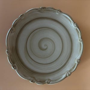 May include: A white ceramic plate with a swirl design in shades of brown. The plate has a scalloped edge with decorative details.