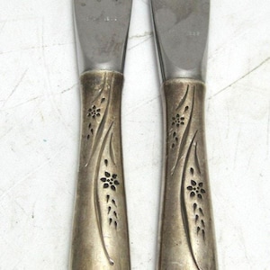 May include: Two silver butter knives. The blades are smooth and curved. The handles are decorated with a floral design. The knives are made of a shiny, metallic material.