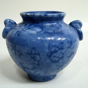 May include: A rounded ceramic vase featuring a vibrant blue glaze with a mottled, cloud-like pattern in lighter shades of blue and white. The vessel has a wide opening, a tapered base, and two small, curved handles attached to the upper sides.