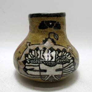 May include: A small, handcrafted ceramic vase with a rounded body and a narrow neck. The vase features a tan background with black and white geometric designs and a black rim. The design includes a stylized leaf and other abstract shapes.