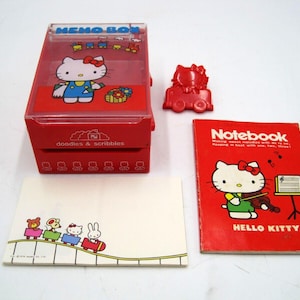 May include: A red Hello Kitty memo box, notepad, and accessories. The memo box has a clear lid and features Hello Kitty. The notepad is red with Hello Kitty playing the violin. A red Hello Kitty car and notepad are also included.