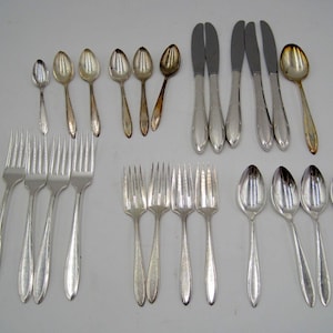 May include: An assortment of vintage silverware, including forks, spoons, and knives, arranged on a light-colored surface. The silverware is primarily silver-toned, with some pieces showing signs of age. The collection includes various sizes and styles of utensils.
