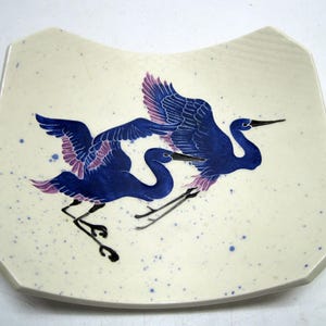 May include: A white ceramic plate with a blue and purple design of two cranes in flight. The plate has a speckled white glaze and a curved shape.
