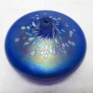 May include: A round, blue glass object with a dark blue opening at the top. The glass has a speckled design of white and light blue shapes. The surface has an iridescent sheen, reflecting colors like purple and gold.