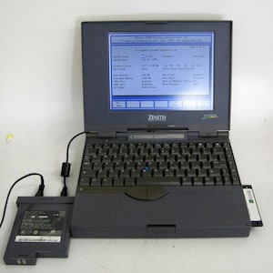 May include: A dark gray Zenith Data Systems Z-STAR EX laptop with a QWERTY keyboard, a blue trackpoint, and a screen displaying Phoenix BIOS setup utility text. A detached rectangular battery pack is connected by a black power cable.