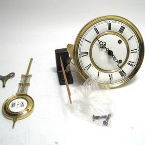 May include: Clock parts including a clock face with black Roman numerals, a pendulum, a key, and other components. The clock face has a gold-colored bezel and black hands. The pendulum has a gold-colored frame and a white face with black lettering.