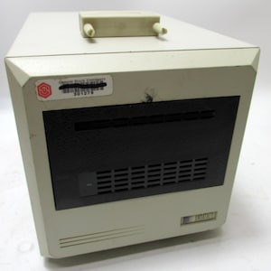 May include: A beige and black computer component with a rectangular shape. It has a black front panel with horizontal vents and a small rectangular slot. A sticker from Oregon State University is visible. A beige plastic piece is on top.