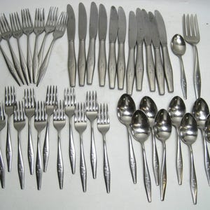 May include: A collection of stainless steel flatware, including forks, knives, and spoons. The silverware has a simple, elegant design with a cross-like pattern on the handles. The set includes multiple forks, knives, and spoons.