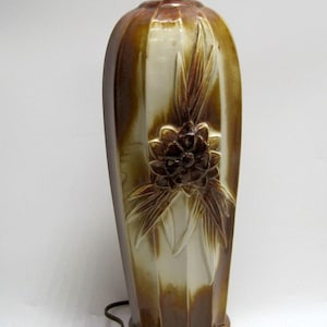 May include: A vintage ceramic table lamp with a brown and cream color scheme. The lamp features a floral design and a brown electrical cord. The lamp's base is a darker brown, and the top has a brass fitting for the lightbulb.