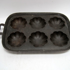 May include: A black cast iron muffin pan with six individual molds shaped like a flower. The pan has two handles on the sides.