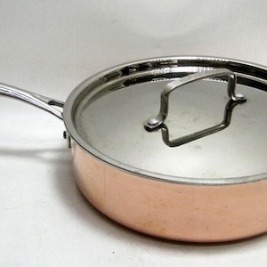 May include: A copper-colored saute pan with a stainless steel lid and handle. The pan has a long, curved handle on one side and a smaller loop handle on the other. The lid has a handle for easy lifting.