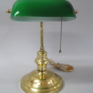 May include: A banker's lamp featuring a polished brass base and a cylindrical stem supporting a curved, emerald green glass shade. The lamp includes a gold-toned pull chain switch hanging from the underside of the shade and a beige power cord.