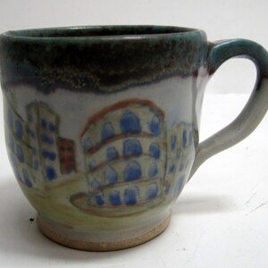 May include: Hand-painted ceramic mug with a cityscape design. The mug features a light blue, white, and brown color scheme, with a dark rim and a curved handle. The artwork depicts buildings with blue arched windows and red roofs.