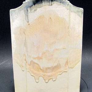 May include: A ceramic vase with a unique, arched top and a cream-colored base. The vase features a layered design with shades of beige, tan, and light blue, creating a textured, artistic appearance. The overall height is approximately 10 inches.